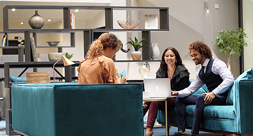 Three people having an informal meeting or conversation in a modern, stylish lounge area with teal couches and shelving units in the background.