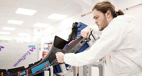 A man in a white cleanroom suit meticulously works on assembling a black composite material structure in a bright manufacturing environment, with "EcoTherm" text visible in the background.
