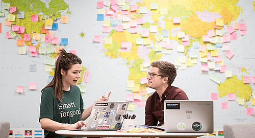 Two colleagues working and discussing at a desk with laptops, positioned in front of a large wall map covered in colourful sticky notes.