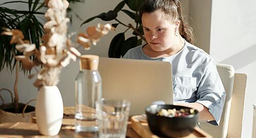 A young person with Down syndrome sits at a sunny dining table, intently using a laptop computer, with house plants, a water bottle, and a bowl of food visible in the foreground.