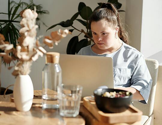 A young person with Down syndrome sits at a sunny dining table, intently using a laptop computer, with house plants, a water bottle, and a bowl of food visible in the foreground.