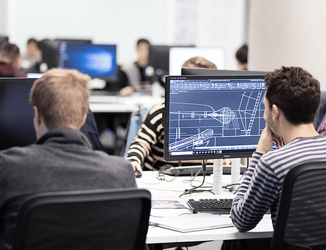 Two people work in a busy computer lab; the monitor in the foreground displays detailed technical drawings, likely using computer-aided design (CAD) software for an engineering or design project.