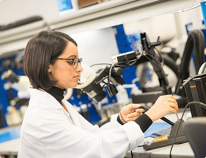 An engineer wearing a white lab coat and glasses uses test probes to work on a piece of electronic equipment or circuit board at a lab bench, likely at a BAE Systems facility.
