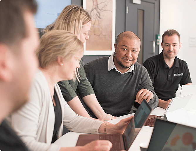 A diverse group of colleagues collaborate around a desk with laptops in a bright office, smiling at each other.