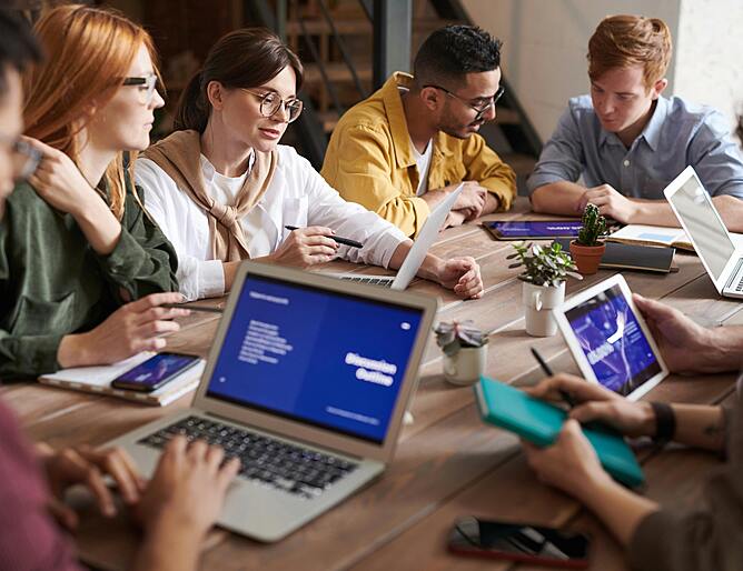 A group of diverse professionals collaborating around a meeting table with laptops and a tablet.
