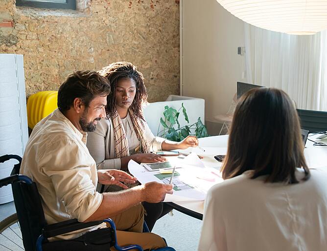 Three people in an office meeting, including a man in a wheelchair, reviewing documents together at a desk.